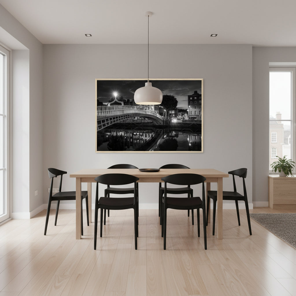Statement black and white photograph of Ha’penny Bridge and Dublin lights reflected in the Liffey, framed in a natural wood frame, 60x90 cm. Stylish dining area with wooden table and black chairs, 60x90 cm Ha’penny Bridge night-time black and white print in natural wood frame on wall, soft pendant light above table, subtle reflections on floor, realistic photography wall art mockup.