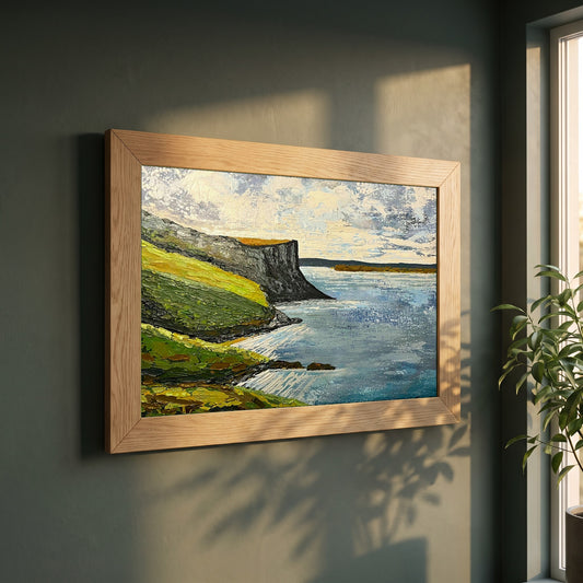 Murlough Bay & Fair Head framed print in a natural oak frame on a dark green wall beside a window, dappled plant shadow