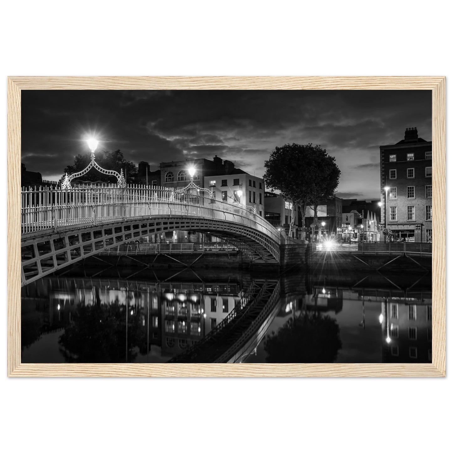  Black and white framed wall art print of the Ha'penny Bridge in Dublin at night. The bridge's historic architecture is beautifully highlighted against the dark sky, with reflections shimmering in the River Liffey below. Ideal for adding elegance to any room or office.