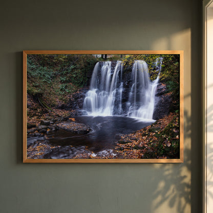 Glenariff Waterfall framed print in a natural oak frame on a dark green wall beside a window, dappled plant shadow