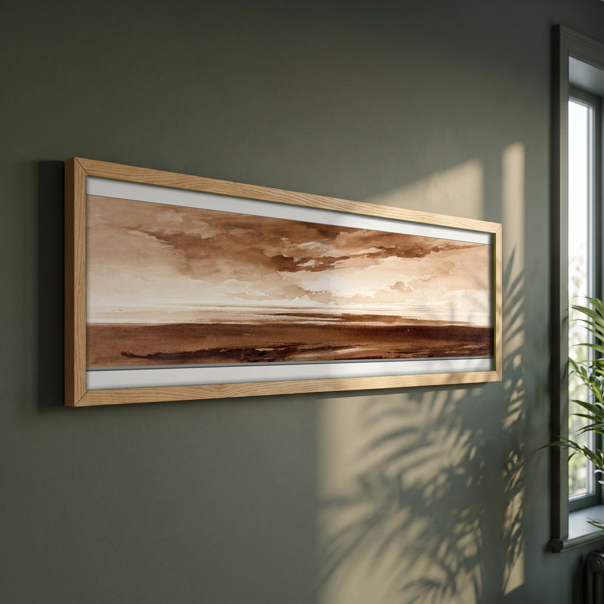 Panorama of the Coast at Sunset framed print in a natural oak frame on a dark green wall beside a window, dappled plant shadow