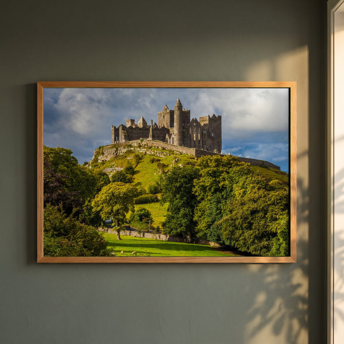 Rock of Cashel framed print in a natural oak frame on a dark green wall beside a window, dappled plant shadow