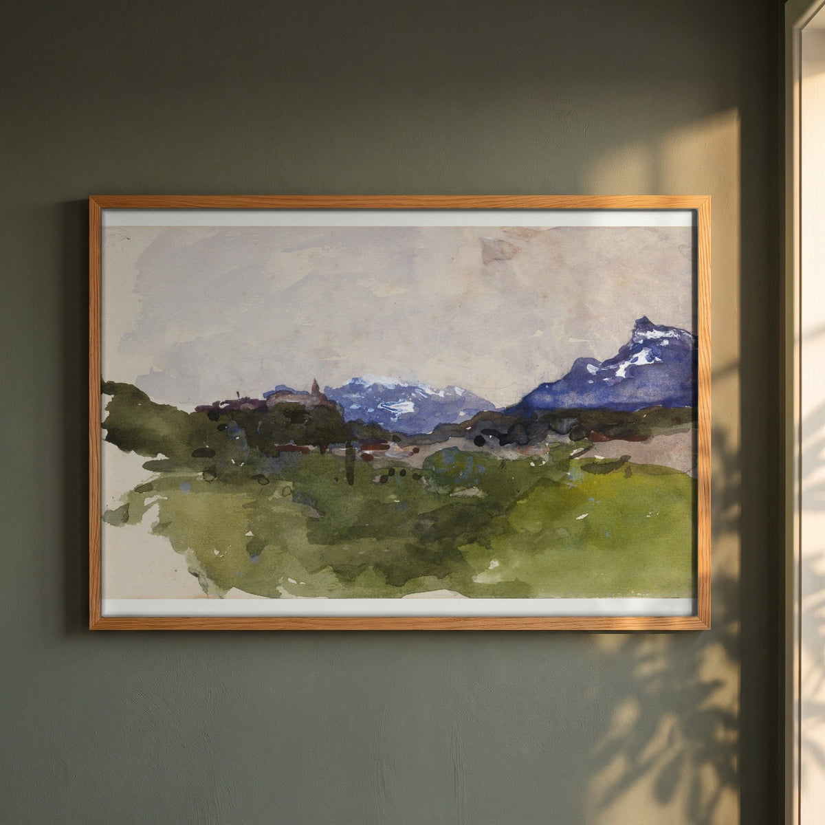The Pyrenees Mountains framed print in a natural oak frame on a dark green wall beside a window, dappled plant shadow